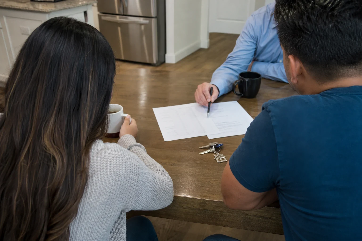 Agent in a blue shirt points a pen at papers on a wooden kitchen table while a couple, seen from behind, looks on beside coffee mugs and a set of house keys