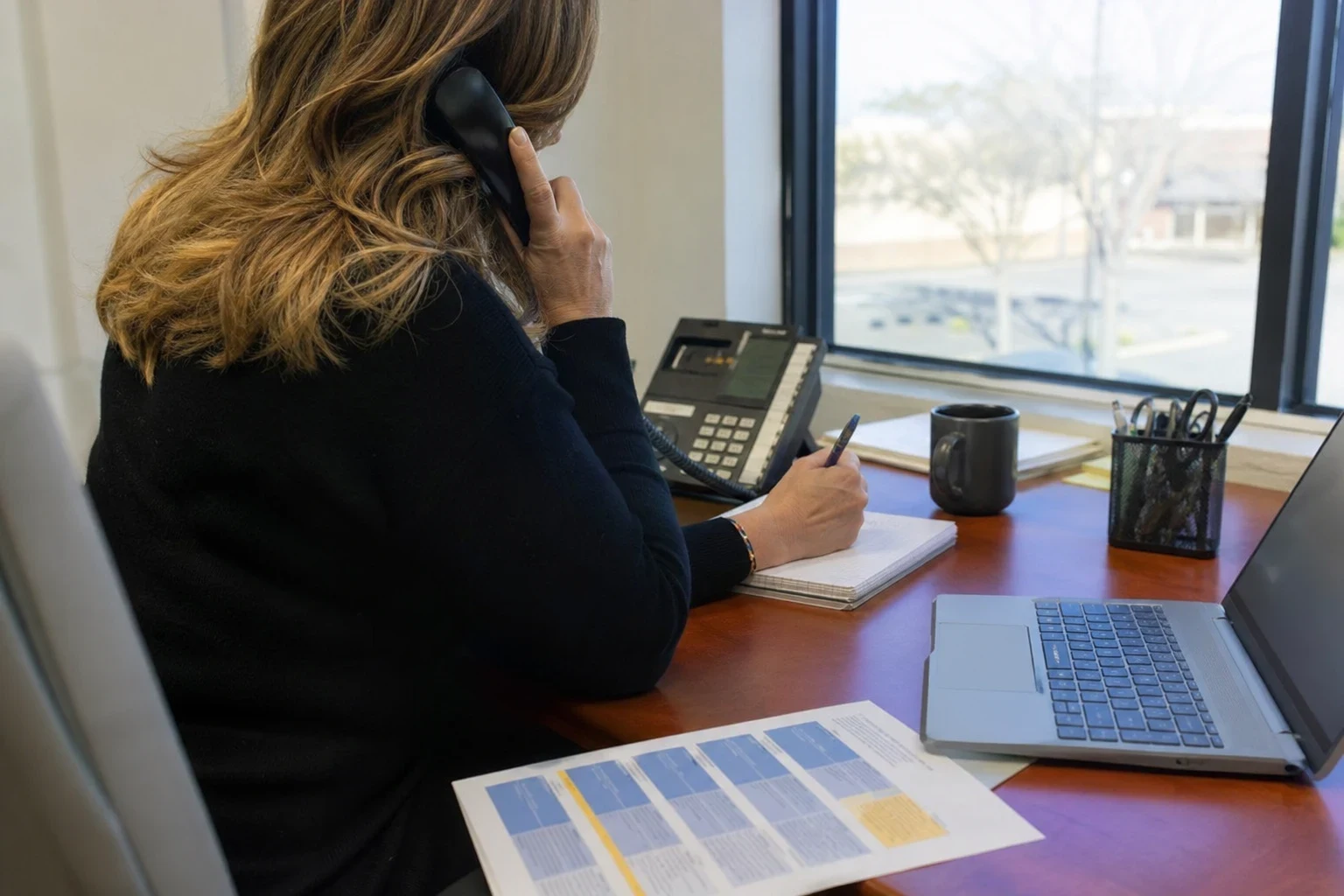 Agent seen from behind on a desk phone, taking notes beside a printed comparison sheet, laptop, mug and pen cup near an office window