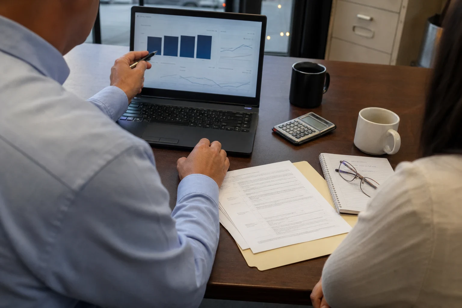 Over-the-shoulder view of a man in a blue button-down pointing a pen at charts on a laptop beside a woman, with papers, a calculator and coffee mugs on an office table